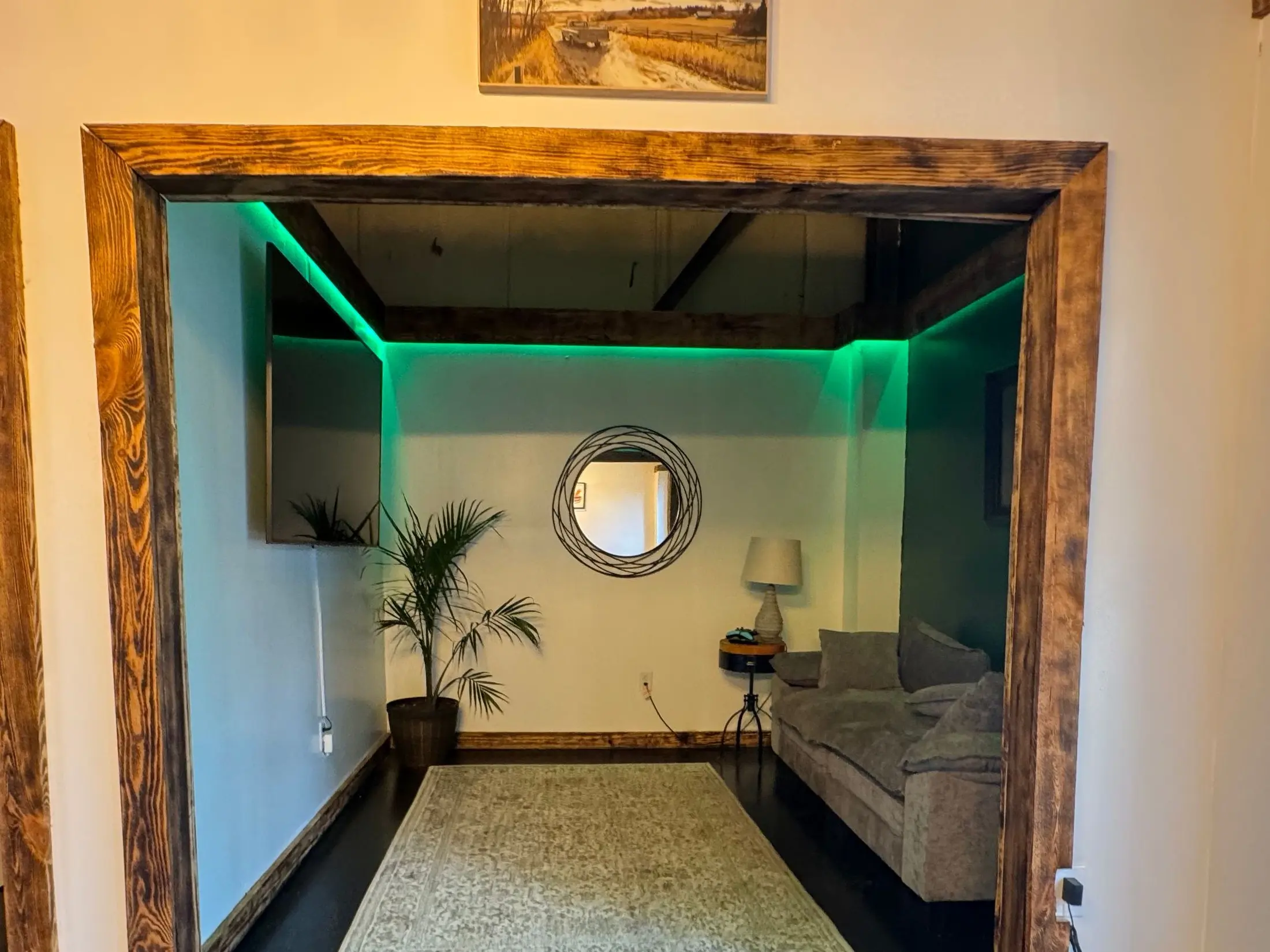 The Whiskey Wrench customer lounge: deep couch, rug, palm and round mirror under green LED cove lighting, framed by burned-pine timber.