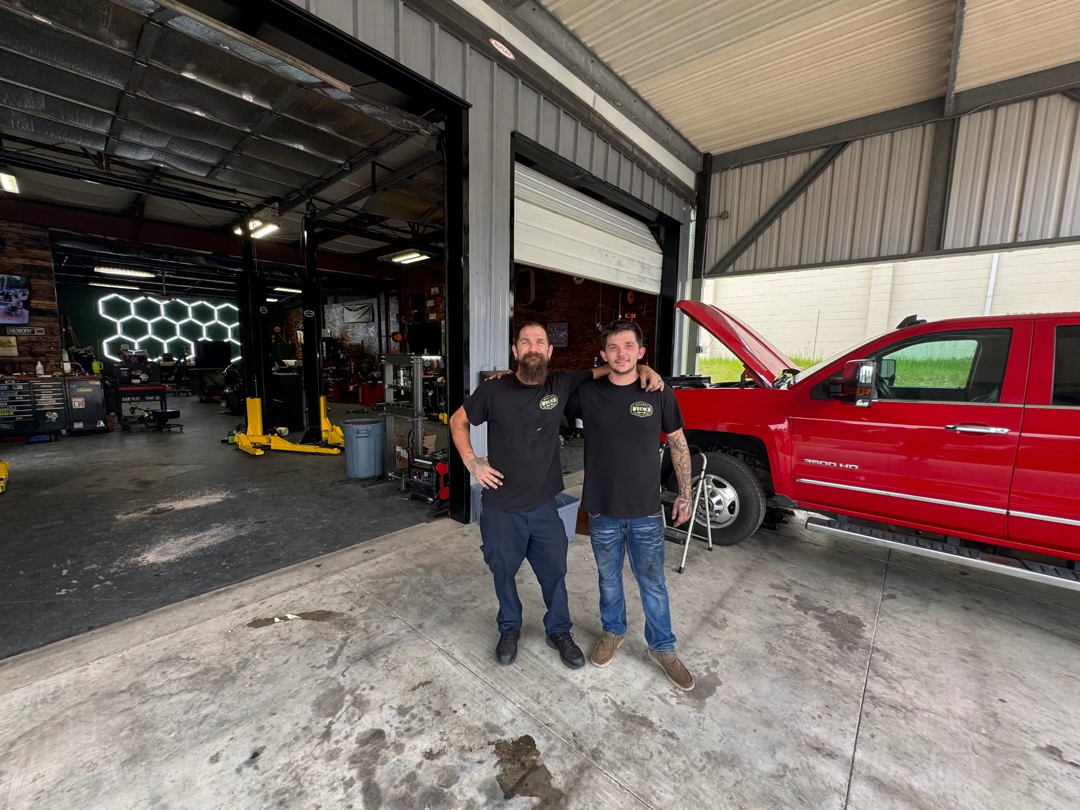 The two Whiskey Wrench mechanics in shop shirts at the open bay door, red Silverado 3500HD behind them.